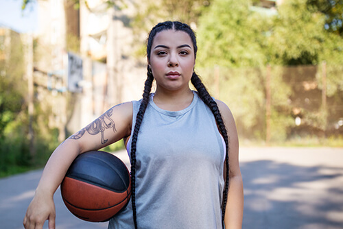 A woman with long braids holds a basketball under her arm.