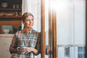 A woman holds a coffee mug and looks out the window.
