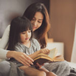 A woman and her daughter sit on a bed reading a book together.