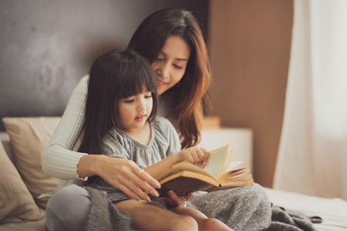 A woman and her daughter sit on a bed reading a book together.