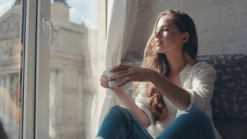 A woman holds a mug and looks out a window.