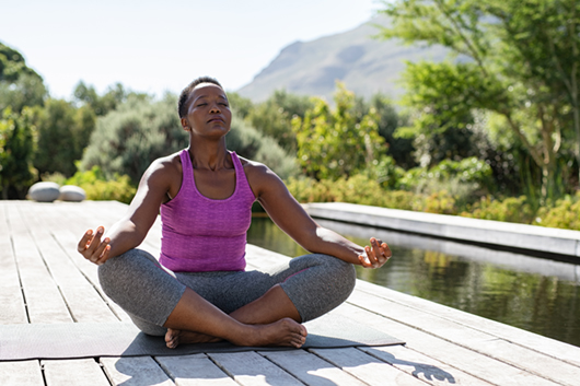 Mature african woman practicing yoga and meditates near swimming pool outdoor. Fitness black lady sitting in lotus pose with closed eyes. African american woman meditating at poolside with copy space.