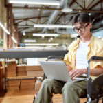 Portrait of young male office worker in a wheelchair looking at the screen of his laptop while performing in co-working space. Disability and handicap concept. Horizontal shot. Selective focus