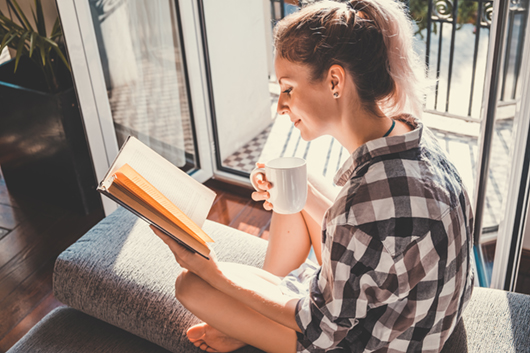 Young pretty woman sitting at opened window drinking coffee and reading a book enjoys of rest