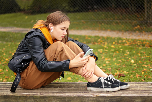 Portrait of modern teenager girl using mobile phone with wireless earphones sitting with her feet on bench outdoors. Child using smartphone.