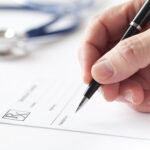 Stock image with a doctor's hand writing a prescription on a Rx blank form. Nearby is seen his stethoscope on the table.