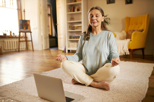 Attractive senior woman sitting on carpet in front of open laptop keeping eyes closed and legs crossed, meditating to nature sounds