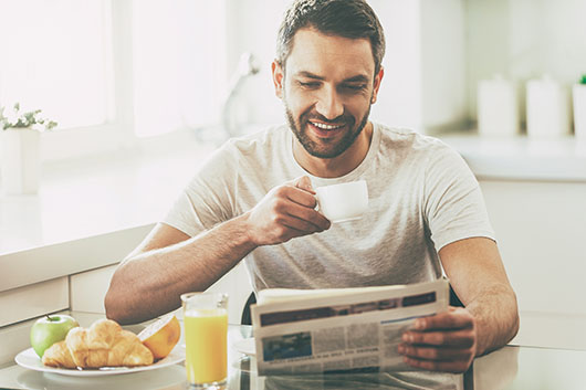 Starting day with good news. Handsome young man reading newspaper and smiling while drinking coffee and having breakfast in the kitchen