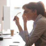 Exhausted female worker sit at office desk take off glasses feel unwell having dizziness or blurry vision, tired woman employee suffer from migraine or headache unable to work. Health problem concept