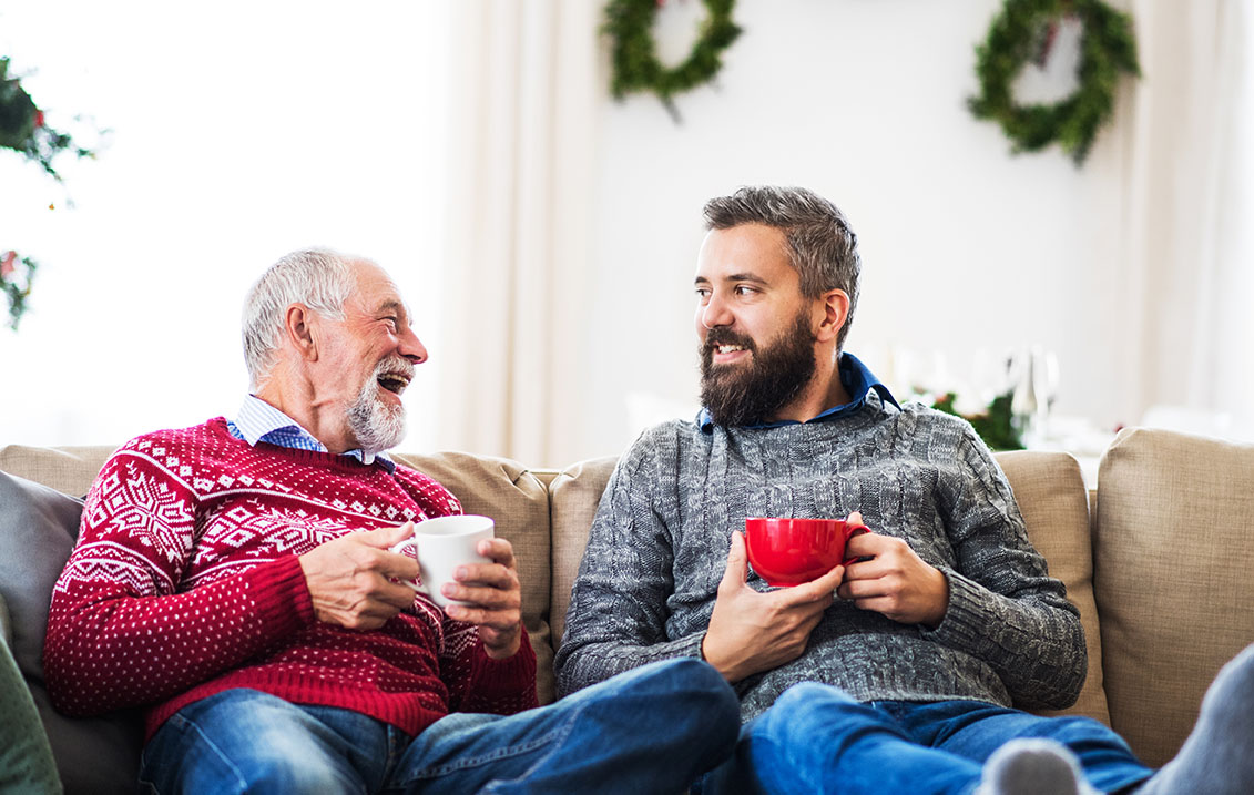 A senior father and adult son sitting on a sofa at home at Christmas time, talking.