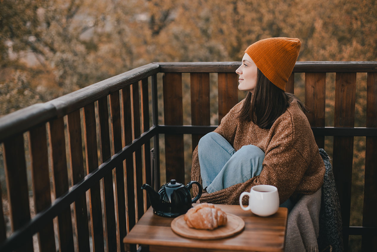 Young woman in knitted sweater and hat drinking tea and eating fresh croissants on cozy balcony of a wooden country house on autumn day.
