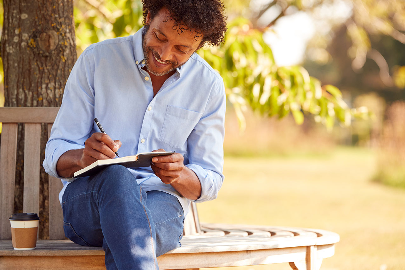 Mature Man With Takeaway Coffee Sitting On Park Bench Under Tree Writing In Notebook