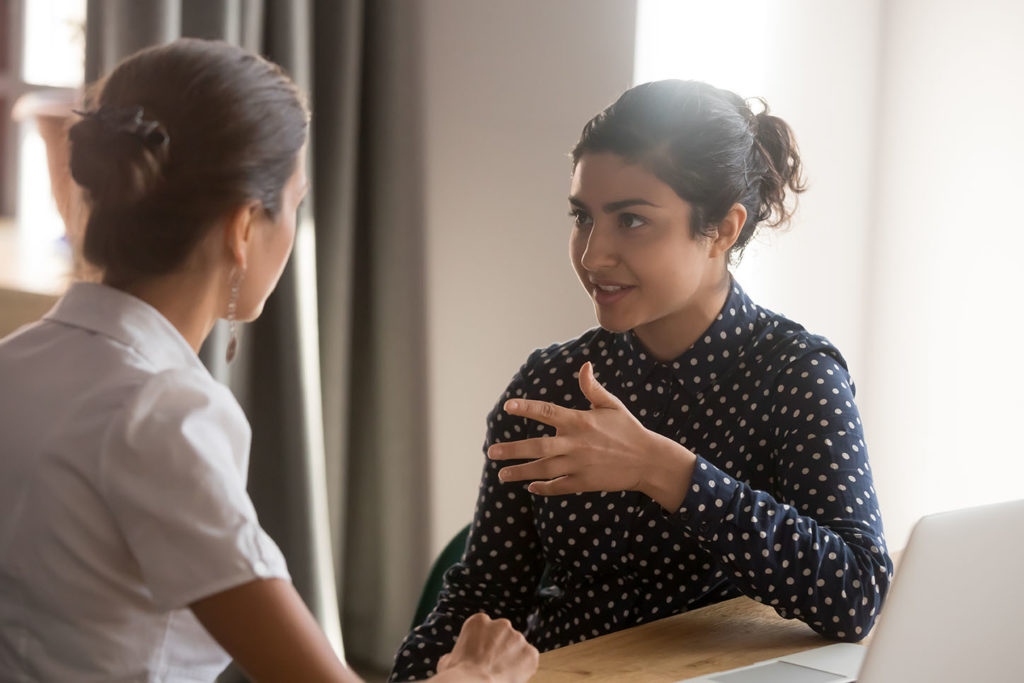 Serious indian mentor teacher worker talk to female colleague teach intern discussing new skills learning sit at work desk, two diverse coworkers work together help cooperate on project in teamwork